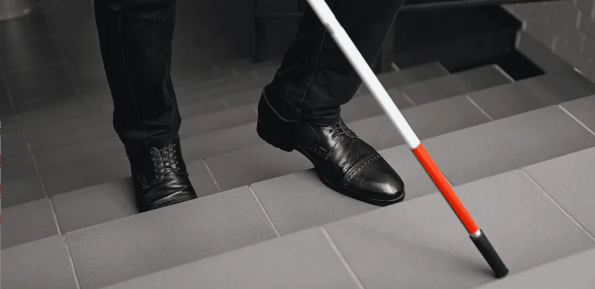 A person wearing black shoes and pants uses a white cane with a red tip to navigate down a tiled staircase.