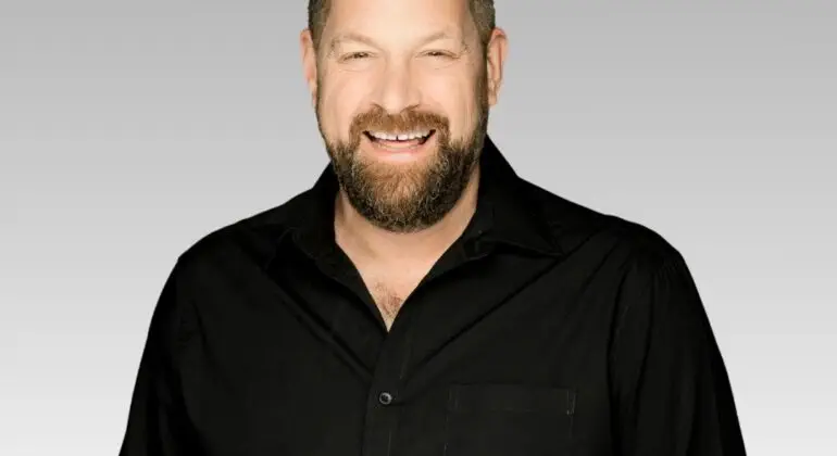 Patrick, a bearded man wearing a black button-up shirt, smiles at the camera in front of a light gray gradient background.