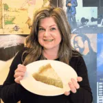 A woman smiles while holding a plate with a slice of pie in front of a wall covered with vintage music album covers. A floating cheesecake slice with caramel sauce and mint is visible in the top right corner.
