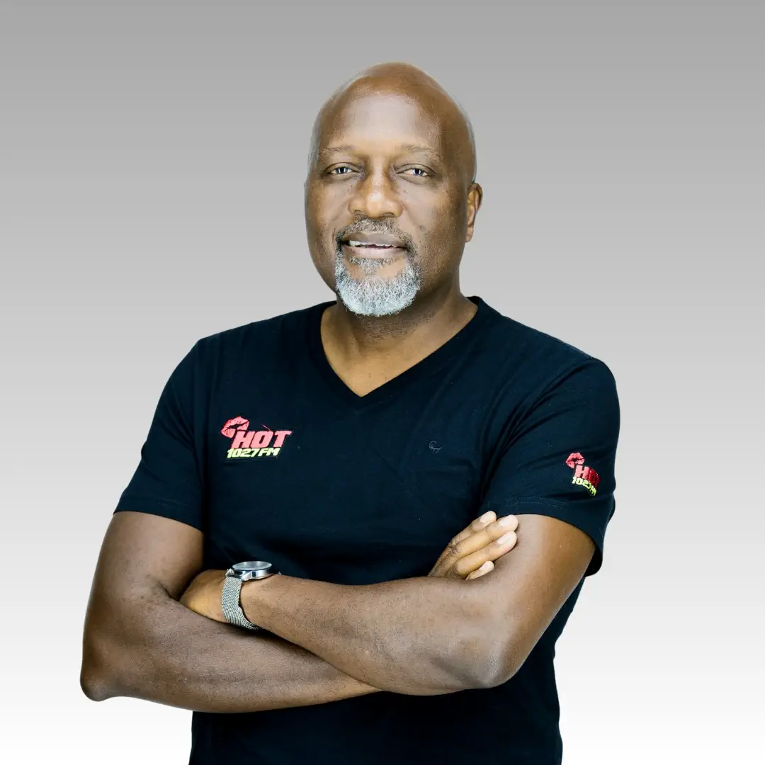 Tony Ndoro with a bald head and gray beard stands with arms crossed, wearing a black t-shirt with HOT logos on the chest and sleeve, and a watch on his left wrist. This could be Tony Ndoro, set against a light gray gradient background.