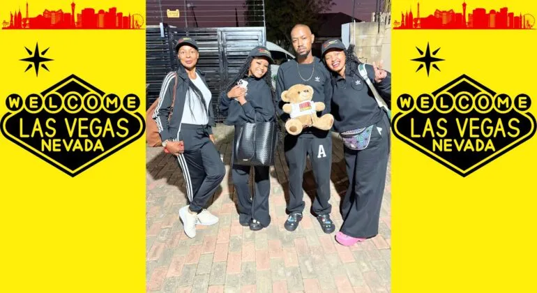 Four people in black outfits pose together, one holding a teddy bear, in front of a brick surface with Welcome to Las Vegas, Nevada signs on a bright yellow background on both sides.