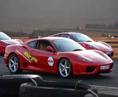 A red sports car with HOT 102.7 FM decals is parked on a racetrack, with another red sports car and a scenic landscape visible in the background. Tires are stacked in the foreground.
