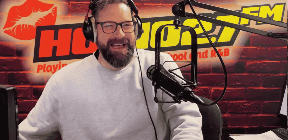 A bearded man wearing glasses and a light gray sweater speaks into a microphone in a radio studio with a red brick wall, HOT 96.7 FM logo, and a red lips graphic in the background.