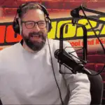 A bearded man wearing glasses and a light gray sweater speaks into a microphone in a radio studio with a red brick wall, HOT 96.7 FM logo, and a red lips graphic in the background.