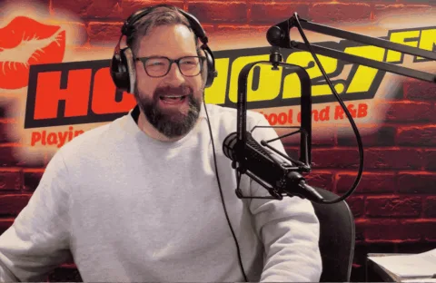 A bearded man wearing glasses and a light gray sweater speaks into a microphone in a radio studio with a red brick wall, HOT 96.7 FM logo, and a red lips graphic in the background.