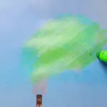 A hand uses a green marker to draw over smoke coming from a factory chimney, making it look like tree foliage against a blue sky.