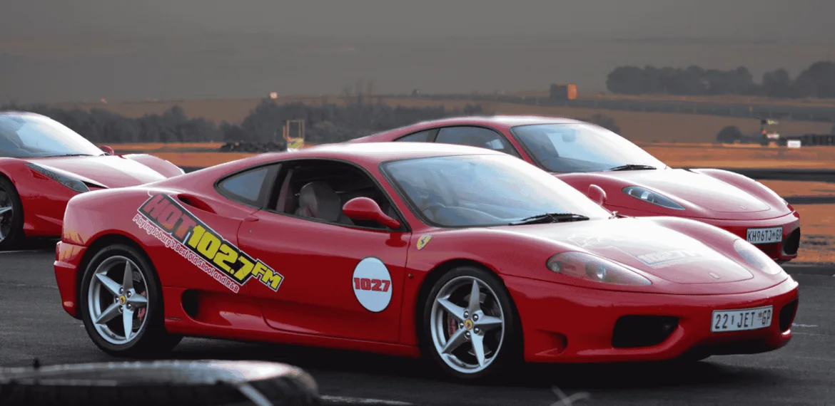 A red Ferrari sports car with 102.7 FM decals parked on a racetrack, with another red Ferrari and open landscape in the background under a cloudy sky.