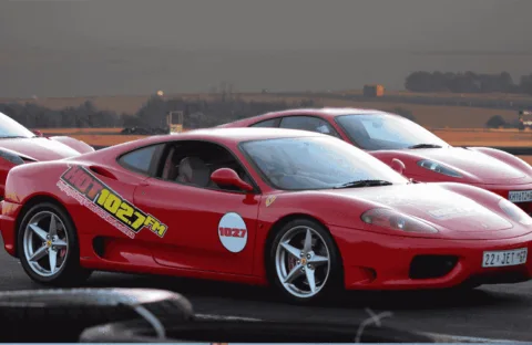 A red Ferrari sports car with 102.7 FM decals parked on a racetrack, with another red Ferrari and open landscape in the background under a cloudy sky.