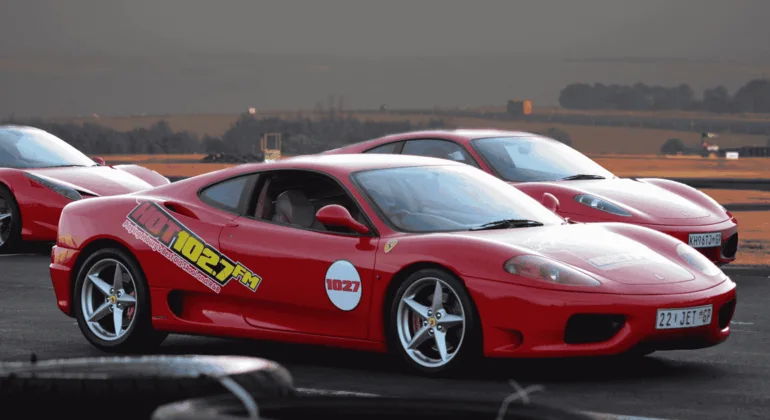 A red Ferrari sports car with 102.7 FM decals parked on a racetrack, with another red Ferrari and open landscape in the background under a cloudy sky.
