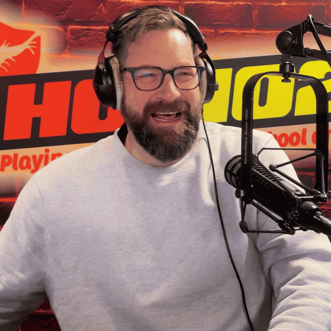 A man with glasses, a beard, and headphones is smiling while speaking into a microphone in a radio studio. A colorful sign with bold red and yellow letters is visible in the background.